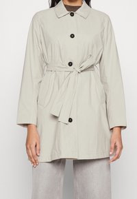 Trench-coat gris clair avec une ceinture à la taille, manches longues et boutons noirs. Caractérisé par un col classique et des poches latérales. Texture de tissu lisse.