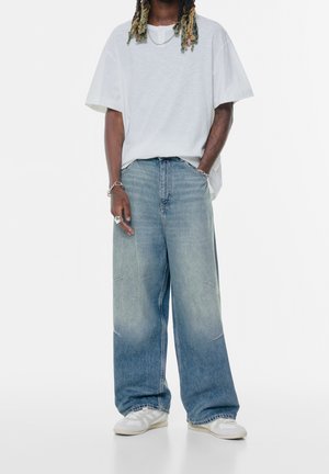 Mand med dreadlocks iført en oversized hvid t-shirt, brede falmede blå jeans, hvide sneakers og sølv smykker mod hvid baggrund.