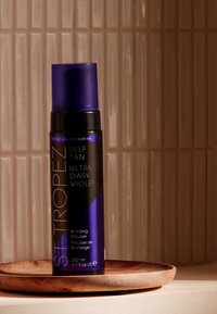 Mousse do samodzielnego opalania w cylindrycznej fioletowej butelce z czarnymi akcentami, oznaczonej jako "Ultra Dark Violet." Prezentowane na okrągłej drewnianej tacy.