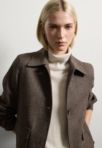 Giovane donna con capelli biondi indossa un giubbotto di lana marrone su un maglione a collo alto bianco, in piedi davanti a una parete bianca semplice.
