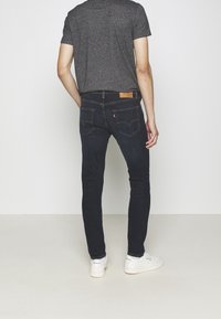 Jeans de mezclilla elásticos de color azul oscuro con un ajuste slim, que presentan costuras contrastantes y una etiqueta de piel marrón en la cintura, combinados con zapatillas blancas.