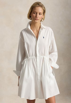 Νεαρή γυναίκα που φοράει λευκό φόρεμα με μακριά μανίκια και ρυθμιζόμενο κορδόνι στη μέση, με μικρό κεντημένο λογότυπο στο στήθος, στέκεται μπροστά από ένα απλό φόντο.