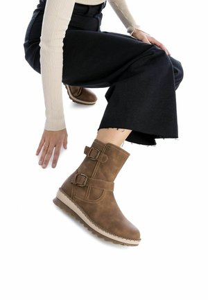 Botas de tobillo marrones hechas de material texturizado, con dos hebillas, una suela de color claro y una punta redondeada. Se ven pantalones amplios negros.