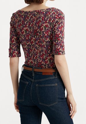 Kvinde iført en lilla blomstermønstret top med albuelange ærmer og mørkeblå jeans med et brunt læderbælte, vist bagfra.