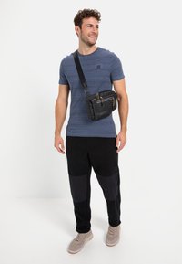 Sort crossbody taske med flere lommer og gyldne lynlåse, båret over en blåstribet shirt og sorte bukser med kontrasterende lapper.
