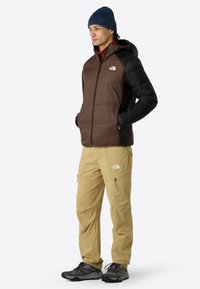 Isolierte braune und schwarze Jacke mit Kapuze, kombiniert mit leichten beigen Hosen und dunklen Schuhen. Verfügt über Logo-Details und Reißverschlusstaschen.