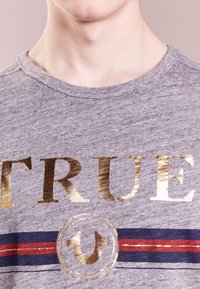 Grå bomullströja med rund hals, prydd med stora guldiga bokstäver som säger "TRUE" och en cirkulär logotyp, accentuerad med röda och blå ränder.