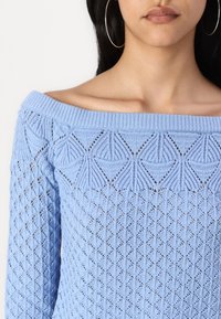 Ljusblå stickad tröja med off-shoulder-design, som har strukturerade diamant- och cirkelmönster i hela plagget.