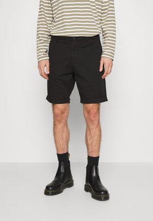 Bermuda nera in cotone con orlo arrotolato, abbinata a una camicia a maniche lunghe a righe. La modella indossa stivali in pelle nera con dettagli gialli.