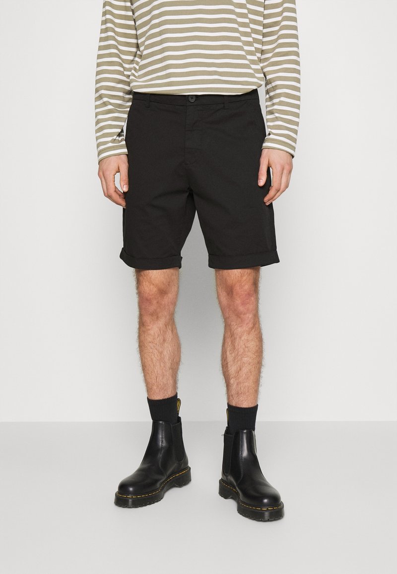 Černé bavlněné šortky s ohrnutým lemem, spárované s pruhovanou dlouhým rukávem. Model má na sobě černé kožené boty s žlutými akcenty.