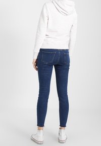 Jeans in denim blu scuro slim fit con vita media, tasche posteriori e orlo sfilacciato, abbinati a una felpa grigia chiara e scarpe da ginnastica bianche.