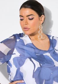 Fließende Bluse aus durchsichtigem Stoff mit blauem Blumenmuster, Rundhalsausschnitt und kurzen Ärmeln. Klare, runde Ohrringe vervollständigen den Look.