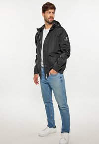 Chaqueta negra impermeable con capucha, cierre de cremallera, puños elásticos y bolsillos laterales. Combinada con jeans de color azul claro y zapatillas blancas.