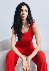 Νεαρή γυναίκα με μακριά σγουρά μαλλιά, φορώντας κόκκινο αμάνικο μπλουζάκι και παντελόνι, καθισμένη σε μια ανοιχτόχρωμη επενδεδυμένη καρέκλα, κοιτάζει προς τα πλάγια.