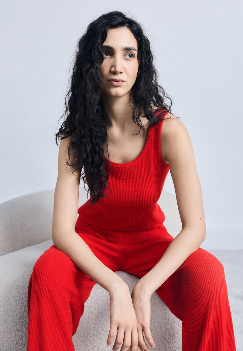 Νεαρή γυναίκα με μακριά σγουρά μαλλιά, φορώντας κόκκινο αμάνικο μπλουζάκι και παντελόνι, καθισμένη σε μια ανοιχτόχρωμη επενδεδυμένη καρέκλα, κοιτάζει προς τα πλάγια.