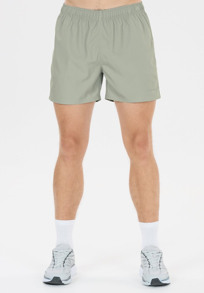 Homme portant un short de sport gris clair à taille élastique, des chaussettes blanches montantes et des chaussures de course grises, debout devant un fond blanc uni.