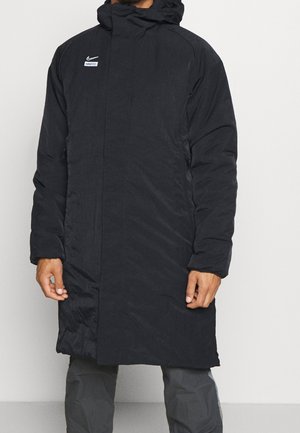 Homme portant un long manteau d'hiver noir Nike FC avec capuche, debout devant un fond clair uni.