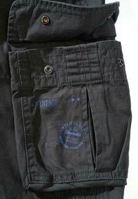 Žep iz črne tkanine z zavihek, s srebrno kovino, vidnimi šivi in modrim tiskanem logotipom z napisom "VINTAGE" ter imenom blagovne znamke.