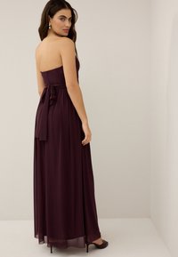 Femme portant une longue robe bustier violet foncé avec un nœud noué dans le dos, debout, regardant par-dessus son épaule dans une pièce neutre.