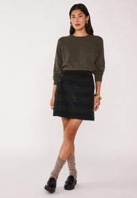 Maglione verde oliva con maniche raglan abbinato a una gonna a quadri in tonalità scure, con dettagli di bottoni e orlo dritto. Calze marroni e scarpe nere.