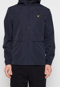 Homme portant une veste légère noire à capuche avec poches avant et un petit logo d’oiseau jaune sur la poitrine gauche.