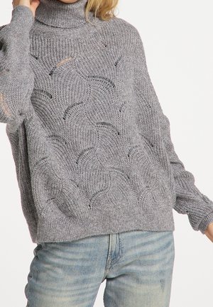 Pull en tricot gris avec un col montant, présentant un motif en vagues texturé, coupe ample et poignets et ourlet côtelés. Jeans en denim visibles en dessous.
