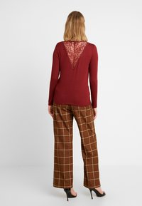 Långärmad bordeauxfärgad topp med en spetsdetalj på V-backen, parat med bruna rutiga vida byxor och svarta klacksandaler.