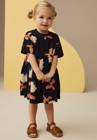Abito nero con motivi a fiocco sovradimensionato in rosso e bianco, tessuto testurizzato, maniche corte, abbinato a sandali marroni su una bambina.