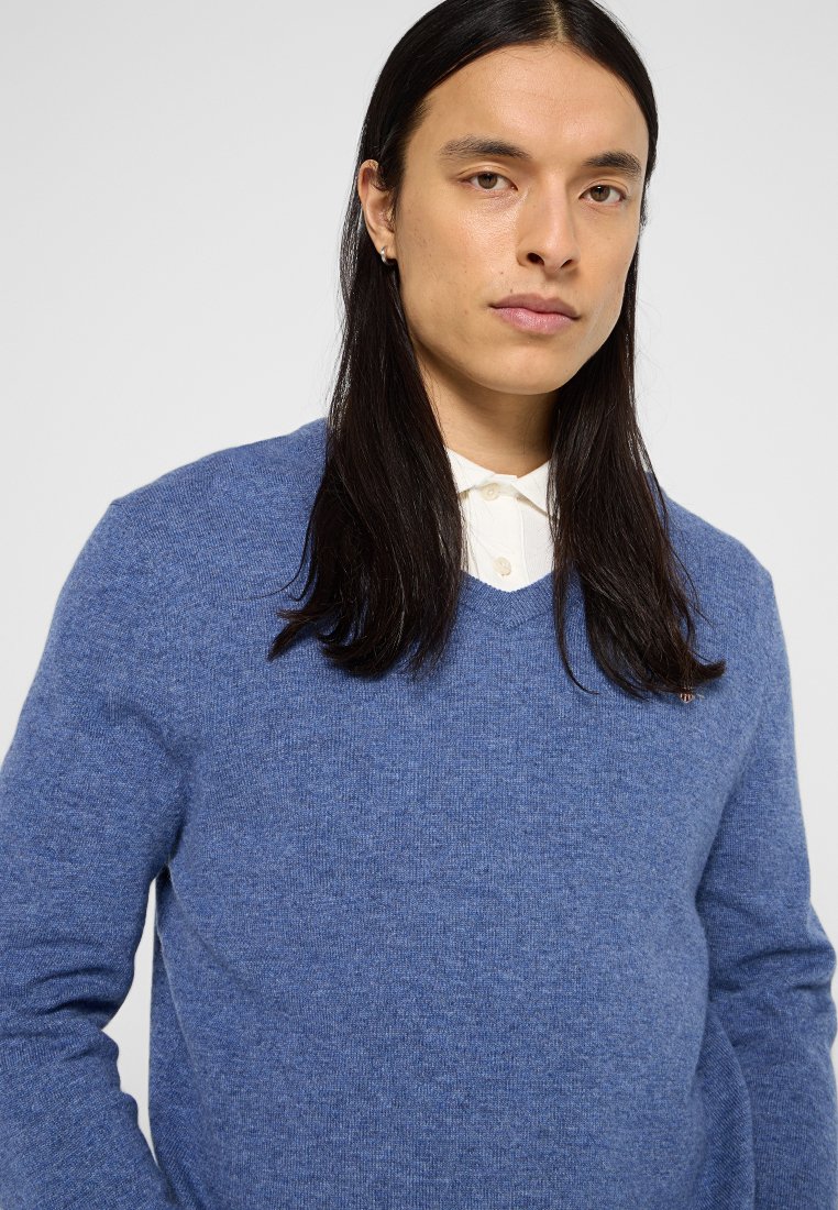 Niebieski sweter wykonany na drutach z dekoltem w kształcie litery V, wykonany z miękkiego, fakturowanego materiału. Posiada długie rękawy i warstwę białej koszuli pod spodem.