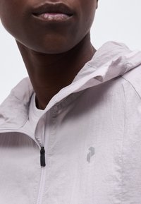 Κοντινό πλάνο ενός ατόμου που φορά γκρι ανοιχτό μπουφάν με φερμουάρ, με μικρό γεωμετρικό λογότυπο στο στήθος και ανεβασμένο γιακά.