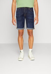 Shorts in denim per uomo di colore blu scuro, con orlo arrotolato, design a cinque tasche e una texture leggermente sbiadita, abbinati a scarpe da ginnastica bianche alte.