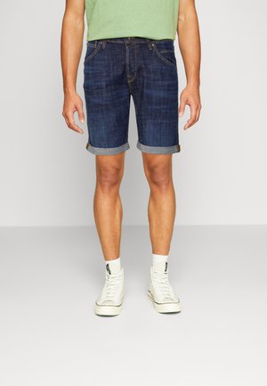 Bermudas de mezclilla para hombre en azul oscuro, con dobladillo enrollado, diseño de cinco bolsillos y una sutil textura desgastada, combinadas con zapatillas altas blancas.