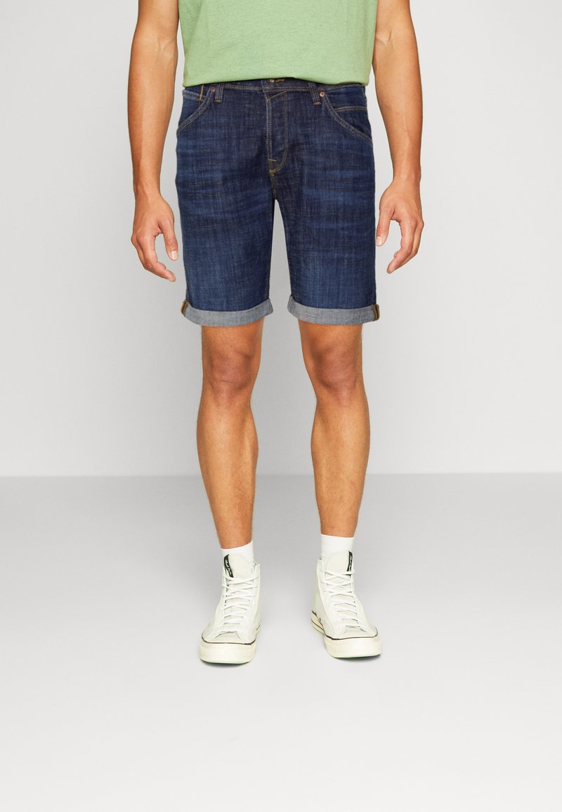 Shorts in denim per uomo di colore blu scuro, con orlo arrotolato, design a cinque tasche e una texture leggermente sbiadita, abbinati a scarpe da ginnastica bianche alte.