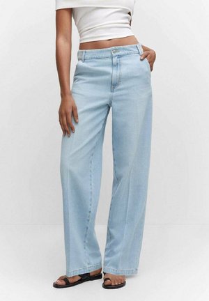 Frau trägt hellblaue Jeans mit weitem Bein, weißes kurz geschnittenes Top und schwarze flache Sandalen, eine Hand in der Tasche, vor einfarbigem Hintergrund.