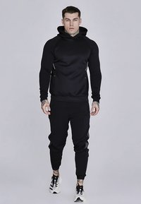 Fekete sportos melegítőfelső kapucnival, cipzáras zsebekkel és testhezálló jogger nadrággal. Az anyag sima, fényes felületű.