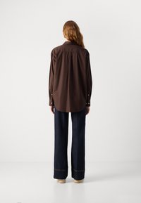 Brown long-sleeve shirt with a curved hem and button cuffs, paired with dark blue wide-leg jeans and light-colored shoes.
