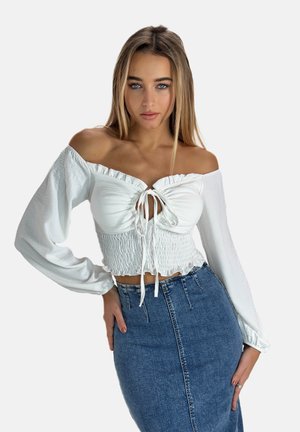 Weißes Off-Shoulder-Oberteil mit Bindung, geraffter Textur und ausladenden langen Ärmeln. Kombiniert mit einem figurnahen, dunkelblauen Jeansrock.