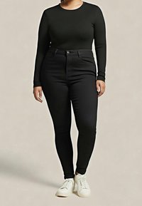 Personne portant un haut à manches longues noir et un jean skinny noir taille haute avec des baskets blanches, debout devant un fond neutre.