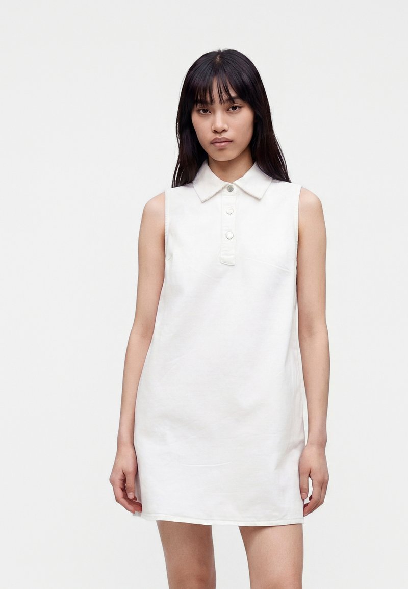 Jeune femme aux longs cheveux foncés portant une robe blanche sans manches avec col et trois boutons sur le devant.