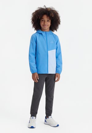 Παιδί με σγουρά μαλλιά που φοράει μπλε μπουφάν με κουκούλα, σκούρο γκρι παντελόνι και λευκά αθλητικά παπούτσια, στέκεται μπροστά σε απλό λευκό φόντο.