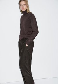 Bruine coltrui van zacht materiaal, gecombineerd met een donkere, op maat gemaakte broek met zijzakken. Eenvoudig, strak ontwerp op een witte achtergrond.