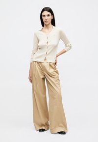 Γυναίκα με μακριά σκούρα μαλλιά, που φοράει μπεζ ριπ ζακέτα και φαρδιά καφέ παντελόνια, στέκεται με το ένα χέρι στη μέση μπροστά σε λευκό φόντο.