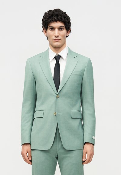 Άνδρας με σγουρά σκούρα μαλλιά, ντυμένος με ανοιχτό πράσινο κοστούμι, λευκό πουκάμισο και μαύρη γραβάτα, στέκεται μπροστά σε απλό ανοιχτό φόντο.