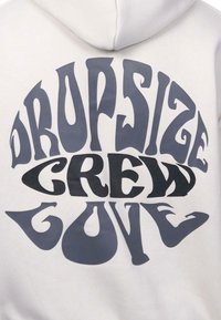 Crewneck sweatshirt in light beige with bold, graphic text in dark grey. The lettering features a curved, psychedelic style and varied shapes.