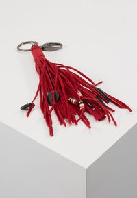 Porte-clés en daim rouge avec un anneau en métal, plusieurs brins en cuir, perles colorées et petits accents métalliques décoratifs sur une surface blanche.