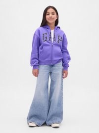 Ragazza in piedi che indossa una felpa viola con zip con la scritta "GAP" e jeans a gamba larga di un blu sbiadito, contro uno sfondo bianco puro.
