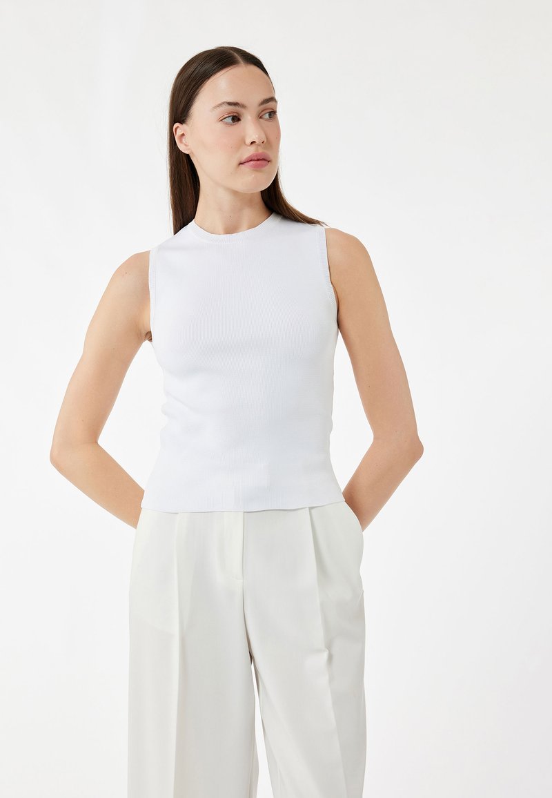 Hvid ribbet ærmeløs top med høj hals, parret med lysfarvede wide-leg bukser. Stoffet ser glat og tætsiddende ud.