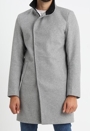 Homme portant un manteau en laine gris clair longueur genou avec des boutons cachés sur le devant et un col intérieur noir, associé à un jean bleu.