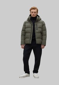 Chaqueta acolchada verde con capucha, cierre frontal y diseño acolchado. Lleva puesta sobre un suéter negro y combinada con pantalones negros y zapatillas blancas.