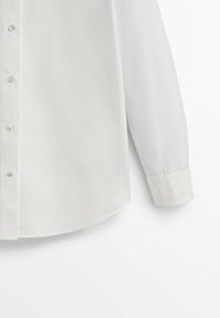 Chemise blanche à manches longues avec manches retroussées et boutons visibles sur la patte de boutonnage avant, sur un fond blanc.